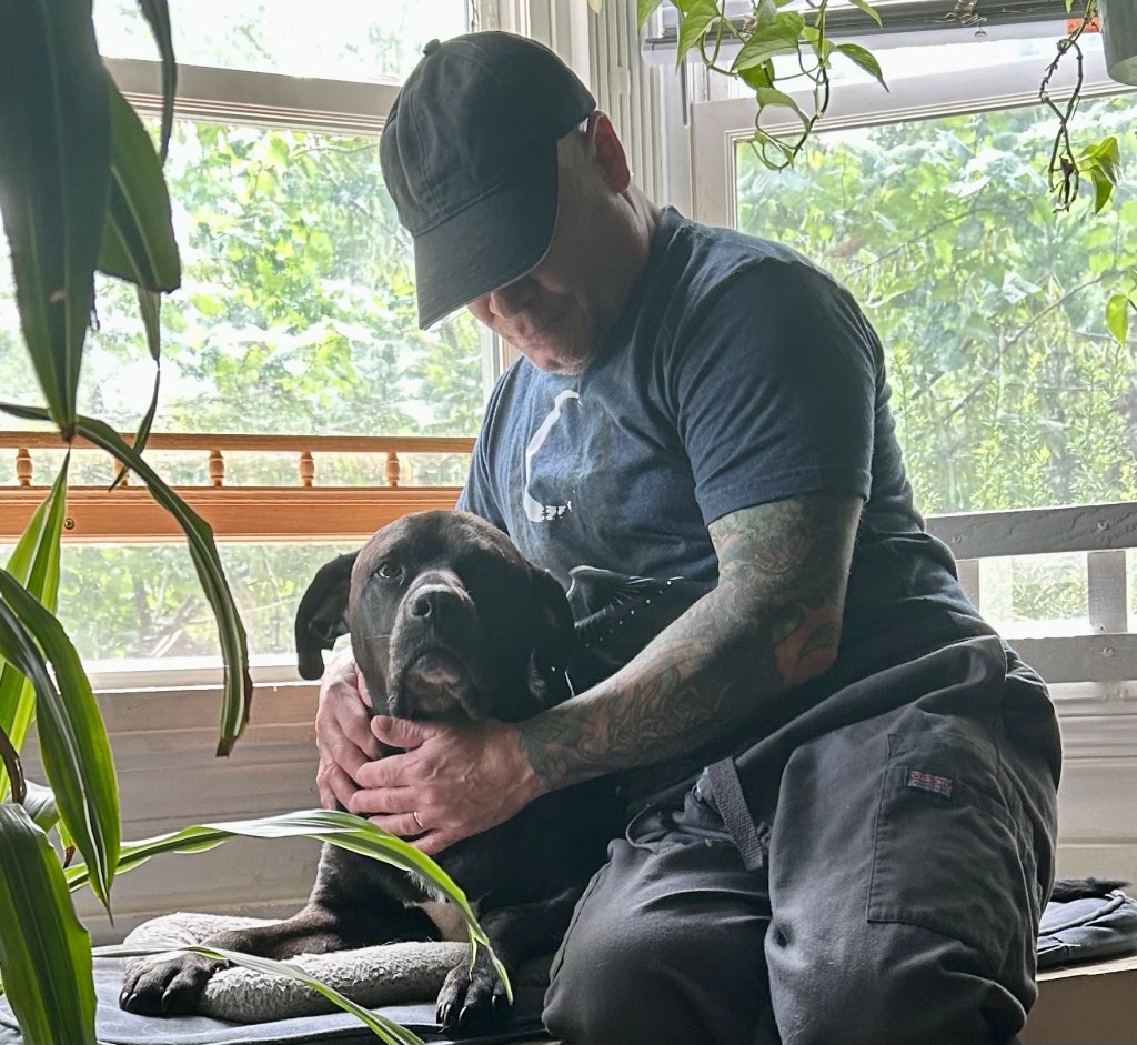 A man wearing a black baseball cap, dark tee shirt, and grey pants sits on a window seat with his arms around a medium-sized dog with white under her chin and on her chest.  The dog is looking straight at the camera, as if warning other dogs in the vicinity not to interrupt her moment.