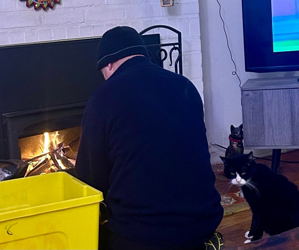 A black and white tuxedo cat watches a man build and light a fire in a fireplace.